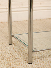Load image into Gallery viewer, Close-up of a modern side table with polished chrome legs and a clear glass shelf, shown on a woven beige rug.
