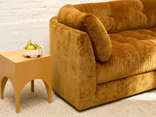 Load image into Gallery viewer, A close-up of a mid-century modern mustard yellow velvet sofa next to a light wood side table with a bowl of pears, set against a white brick wall and beige carpet.
