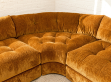 Load image into Gallery viewer, A curved modular sectional sofa upholstered in a rich mustard-gold velvet fabric, featuring deep button tufting across the seat and back cushions, set against a white brick wall background.
