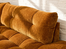 Load image into Gallery viewer, A close-up of a tufted velvet sofa cushion in a warm rust-brown color, set against a white brick wall with soft sunlight and shadows.
