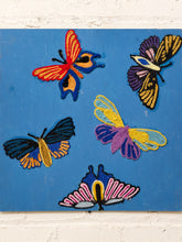 Load image into Gallery viewer, A square blue wall-hanging artwork featuring five colorful embroidered butterflies in red, yellow, purple, black, and pink, displayed against a white brick wall.
