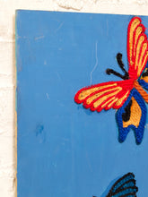 Load image into Gallery viewer, A close-up of a blue wooden wall art panel featuring colorful embroidered butterflies, with one large red, orange, yellow, and blue butterfly on the right and part of another dark blue butterfly at the bottom.
