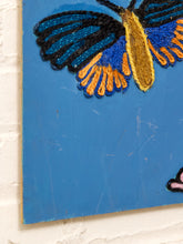 Load image into Gallery viewer, A close-up of a colorful embroidered artwork on a blue rectangular panel, showing a butterfly with blue, black, yellow, and orange details, mounted against a white brick wall.
