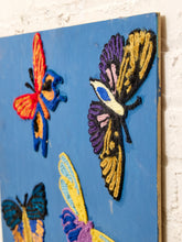 Load image into Gallery viewer, A close-up of a blue wall-hanging or canvas featuring raised embroidered butterflies in bright colors, including red, blue, purple, yellow, and black, mounted against a white textured wall.
