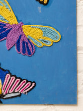 Load image into Gallery viewer, Close-up of a colorful embroidered textile or wall art featuring butterfly and leaf motifs in yellow, purple, blue, pink, and black on a bright blue background, shown against a white brick wall.
