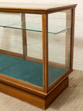 Load image into Gallery viewer, A vintage wooden display case with glass doors and shelves, featuring a teal fabric lining on the bottom, set against a white brick wall and beige carpet.
