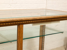 Load image into Gallery viewer, A close-up of a mid-century modern console table featuring a warm medium-brown wood frame, a top glass tabletop, and a lower glass shelf, set against a white brick wall background.
