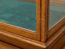 Load image into Gallery viewer, Close-up detail of a wooden display case corner with a warm medium-brown oak frame and a teal-green glass panel.
