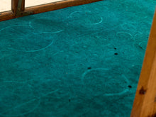 Load image into Gallery viewer, A close-up of a teal green carpeted floor with circular scuff marks and a wooden frame or threshold visible along the top and right edges.
