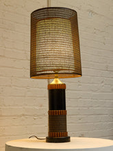 Load image into Gallery viewer, A table lamp with a large cylindrical woven shade, a black and wood base, and a warm glowing light, set against a white brick wall.
