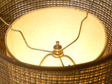 Load image into Gallery viewer, Close-up of a woven lampshade or basket-like light fixture with a warm glowing yellow interior and a brass-colored central finial.

