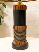 Load image into Gallery viewer, A vintage-style table lamp with a black cylindrical body, fluted bronze-toned metal bands, and a woven rattan or wicker wrap around the middle, set against a white brick wall background.
