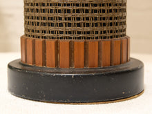 Load image into Gallery viewer, Close-up of a cylindrical decorative object with a black round base, a band of vertical medium-brown wooden slats, and a woven brown basket-like upper section on a light neutral background.
