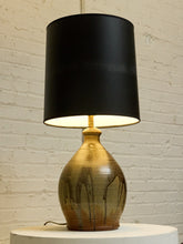 Load image into Gallery viewer, A table lamp with a matte black drum shade and a glazed ceramic base in metallic gold and bronze tones, sitting on a white pedestal against a white brick wall.
