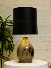 Load image into Gallery viewer, A table lamp with a matte black drum shade and a glossy, gold-toned ceramic base featuring dark brown marbled streaks, displayed on a white pedestal against a white brick wall with a green plant on the left.
