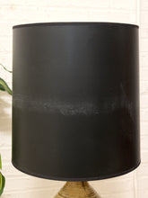 Load image into Gallery viewer, A large black cylindrical lampshade with a slightly distressed or scuffed lower-middle section, mounted on a textured gold-toned base against a white brick wall background.
