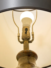 Load image into Gallery viewer, Close-up of a vintage-style table lamp with a brass-toned metal socket, curved metal arms, and a glowing white bulb, framed by a dark lampshade against a warm off-white background.
