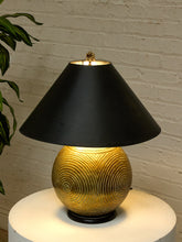 Load image into Gallery viewer, A table lamp with a black conical shade and a textured gold spherical base, illuminated against a white brick wall background.
