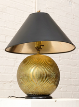 Load image into Gallery viewer, A vintage-style table lamp with a large black conical shade, a gold-toned interior, and a ribbed metallic gold spherical base, set against a white brick wall.
