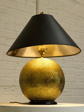 Load image into Gallery viewer, A table lamp with a black conical shade, a black base, and a textured gold globe-shaped body with concentric circular patterns, set against a white brick wall.
