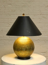 Load image into Gallery viewer, A table lamp with a black conical shade and a textured metallic gold spherical base, shown lit against a white brick wall.
