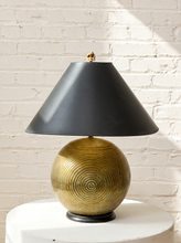 Load image into Gallery viewer, A table lamp with a matte black conical shade and a textured metallic gold spherical base, displayed on a white cylindrical pedestal against a white brick wall.
