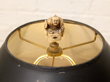 Load image into Gallery viewer, Close-up of a black lampshade with a gold-toned interior and a decorative carved figurine on the center stem, set against a white brick wall background.
