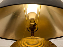 Load image into Gallery viewer, Close-up of a vintage-style brass lamp with a glowing light bulb, featuring a wide gold-toned shade and a ribbed brass base against a white brick wall.
