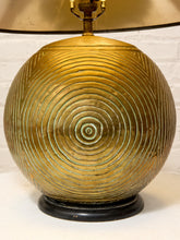 Load image into Gallery viewer, A close-up of a table lamp featuring a spherical, ribbed metallic gold base with concentric circular patterns and a black circular base, with part of a gold lampshade visible at the top.
