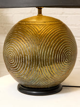Load image into Gallery viewer, A vintage-style table lamp with a large spherical brass-colored shade featuring concentric spiral patterns, a black lamp base, and a black lampshade visible at the top, set against a white brick wall background.
