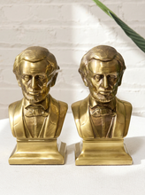 Load image into Gallery viewer, A pair of gold-toned Abraham Lincoln bust bookends on a white surface against a white brick wall background.
