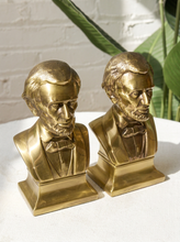 Load image into Gallery viewer, A pair of gold-toned Abraham Lincoln bust bookends on a white surface with a white brick wall and green leaves in the background.
