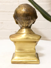 Load image into Gallery viewer, A metallic gold bust sculpture of a person's head and shoulders, shown from the back, set against a white brick wall background with a green leaf in the upper right corner.
