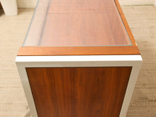 Load image into Gallery viewer, A close-up of a mid-century modern sideboard or cabinet with a warm brown wood grain front, a white frame, and a clear glass top, set on a beige carpeted floor.
