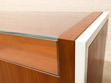 Load image into Gallery viewer, Close-up of a mid-century modern wooden cabinet corner with a glass top, warm brown wood grain, and a white metal frame trim against a light brick wall.
