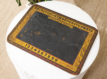 Load image into Gallery viewer, A vintage-style reading board or alphabet board with a black central writing area, yellow border, printed alphabet and numbers, and small decorative illustrations, displayed on a white pedestal.
