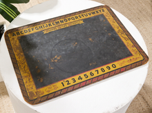 Load image into Gallery viewer, A vintage-style Ouija board with a black central plane, yellow and brown decorative border, alphabet, numbers, and planchette, placed on a white round table.

