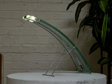 Load image into Gallery viewer, A modern curved glass table lamp with a clear green-tinted glass arc and a silver metal support stand, displayed on a white pedestal against a white brick wall with a green plant in the background.
