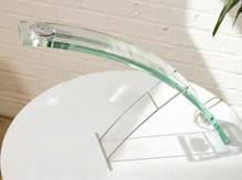 Load image into Gallery viewer, A modern curved glass shelf with a silver metal wire support stand, displayed on a round white table against a white brick wall.

