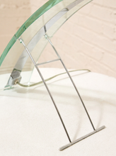 Load image into Gallery viewer, A close-up of a modern glass table or shelf with a curved clear glass top supported by a slim silver metal frame, resting on a white textured surface against a light brick wall.
