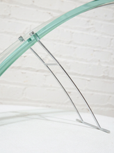 Load image into Gallery viewer, A close-up of a modern curved glass table featuring a clear glass top with a mint green tint and a slim, multi-strand chrome metal base, set against a white brick wall background.
