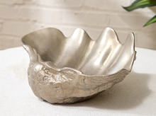 Load image into Gallery viewer, A metallic silver bowl with a textured, organic shell-like shape, resting on a white fabric surface against a light brick wall background.
