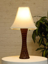 Load image into Gallery viewer, A table lamp with a textured dark brown sculptural base and a warm white fabric shade, displayed on a white round pedestal against a white brick wall with a green plant in the background.
