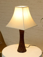 Load image into Gallery viewer, A table lamp with a warm white fabric drum shade and a dark brown textured, scalloped ceramic base, shown lit on a white round table against a white brick wall.
