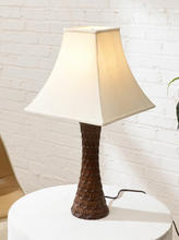Load image into Gallery viewer, A table lamp with a textured dark brown base and a cream-colored fabric shade, illuminated and sitting on a white round table against a white brick wall.
