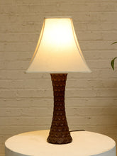Load image into Gallery viewer, A table lamp with a textured dark brown ceramic base and a warm white fabric shade, illuminated against a white brick wall backdrop.
