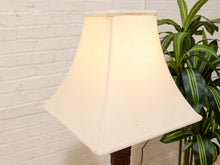 Load image into Gallery viewer, A cream-colored fabric lampshade on a lamp stand, positioned next to a green variegated plant in front of a white brick wall.

