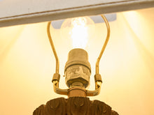 Load image into Gallery viewer, A close-up shot of a vintage-style table lamp with a glowing light bulb, brass-colored metal arms, and a carved wooden base against a warm, illuminated background.

