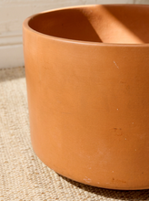 Load image into Gallery viewer, A large terracotta-colored cylindrical planter pot with a matte finish, shown in a close-up shot resting on a woven jute rug.
