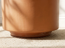 Load image into Gallery viewer, Close-up of a terracotta-colored ceramic planter pot resting on a textured beige carpet with soft natural shadows cast across the surface.
